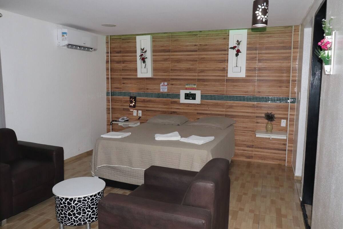A hotel room with a bed, two armchairs, a small table, a wall-mounted phone, and a wall-mounted air conditioner.