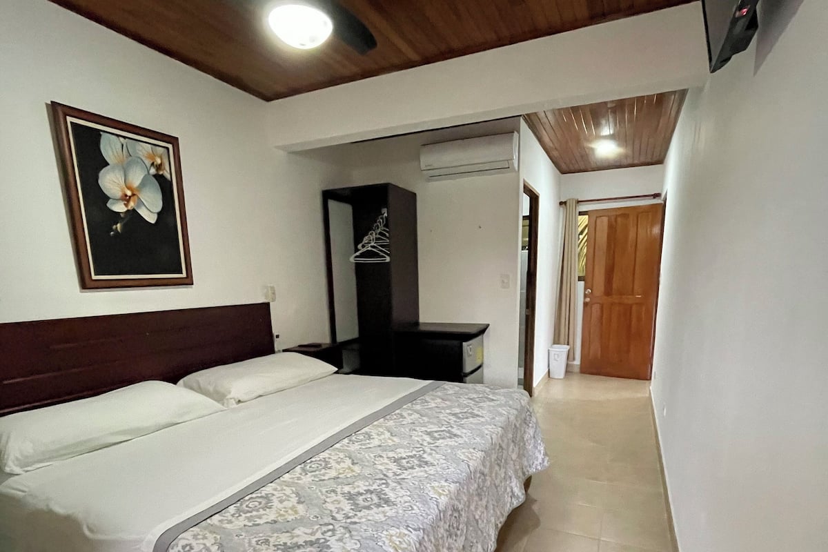 A hotel room with a large bed, a ceiling fan, a framed picture on the wall, and a wooden door in the background.
