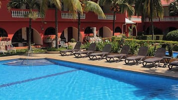 A swimming pool with lounge chairs and palm trees, surrounded by a red building with balconies.