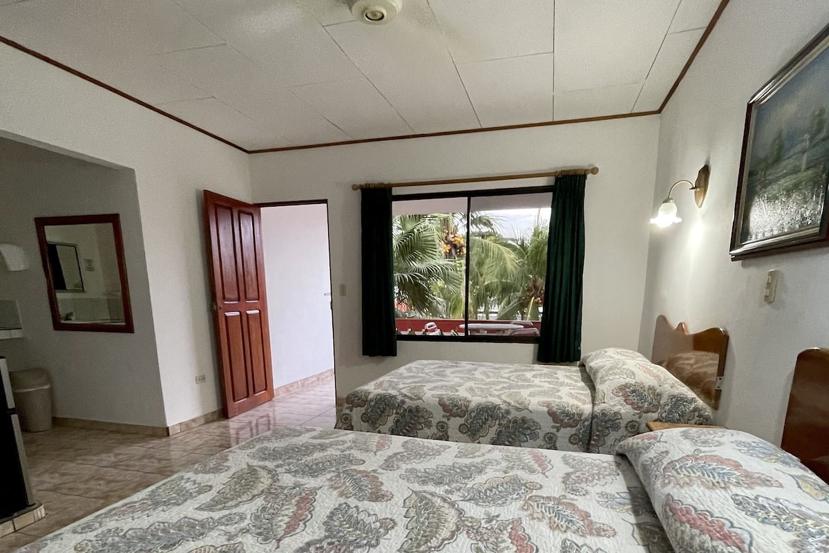 A bedroom with two beds, a ceiling fan, a window with a view of palm trees, and a painting on the wall.