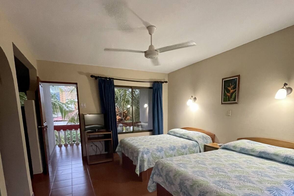 A hotel room with two beds, a ceiling fan, a window with curtains, and a view of the outside.