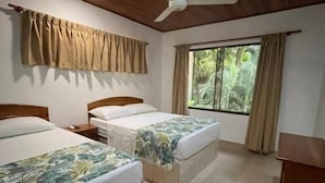 Une chambre avec deux lits, un ventilateur de plafond, une fenêtre avec des rideaux et une table en bois.