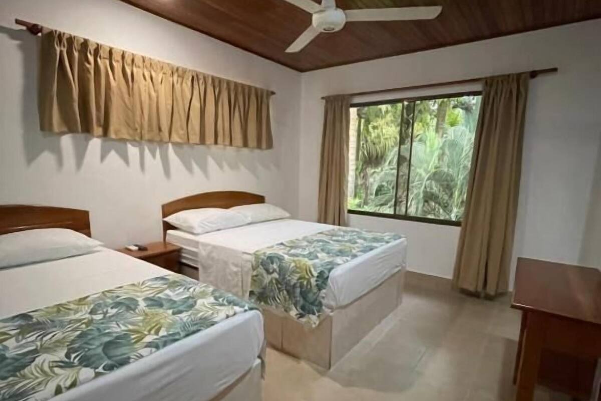 A room with two beds, a ceiling fan, a window with curtains, and a wooden table.