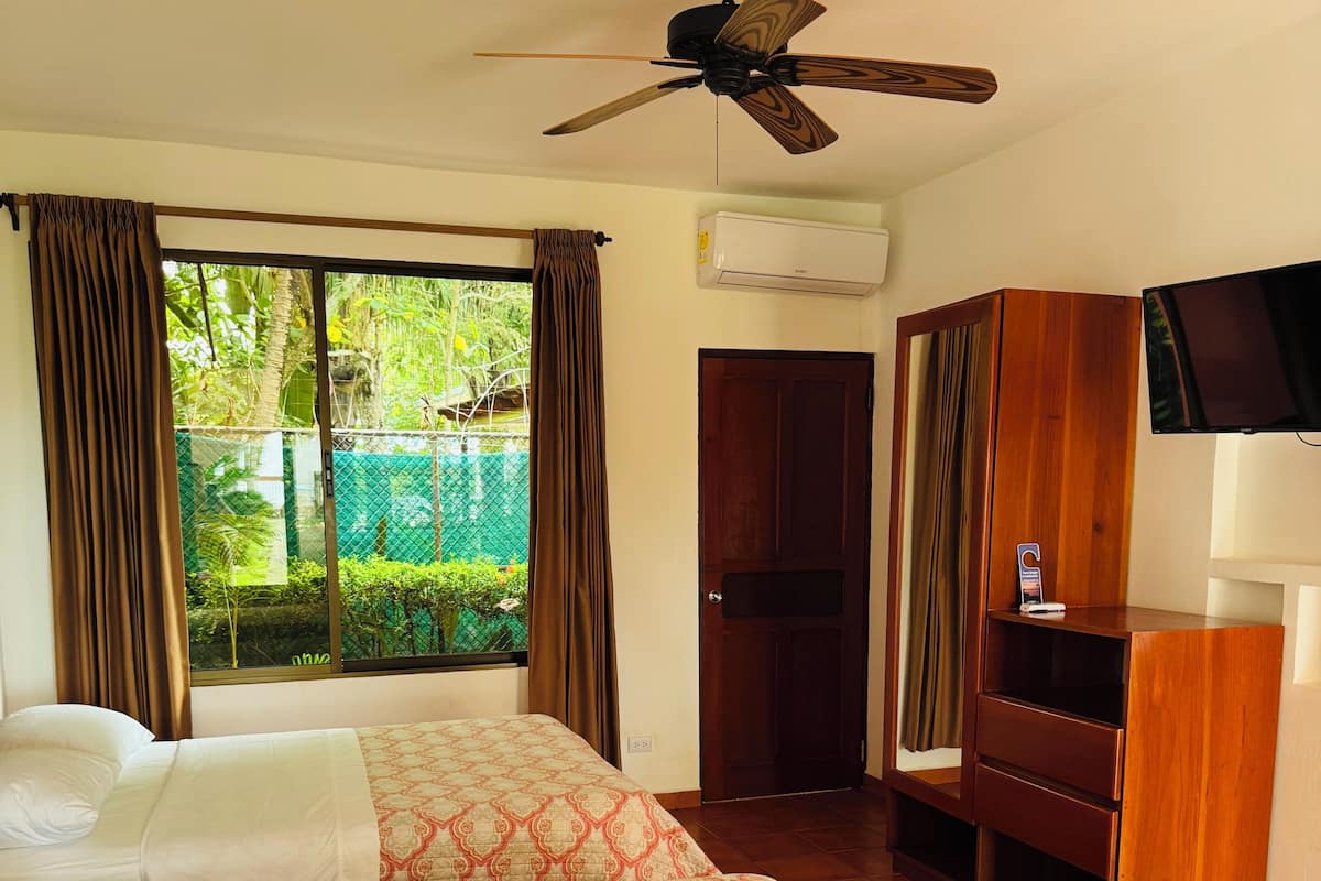 A hotel room with two beds, a ceiling fan, a TV, and a view of greenery outside.