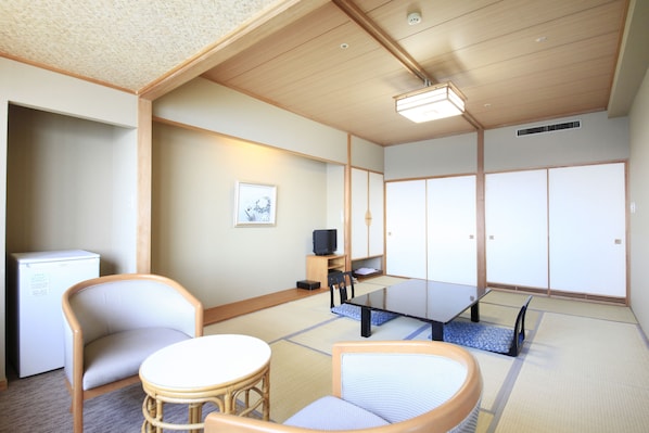Una habitación japonesa tradicional con suelo de tatami, una mesa baja, dos sillas de tatami, un televisor pequeño y un refrigerador.