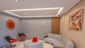 A modern living room with a sectional sofa, two armchairs, a red ottoman, and a wall-mounted circular light fixture. - Hub Hotel (Teresina)