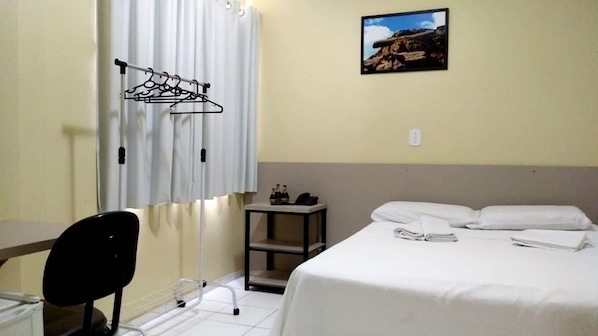 A hotel room with a bed, a desk, a chair, a small table with bottles, and a picture on the wall.