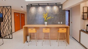 A modern reception area with a wooden reception desk, three yellow chairs, a vase with yellow flowers, and a ceiling light fixture. - Hub Hotel (Teresina)