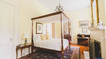 A four-poster bed with a canopy, a nightstand with a lamp, a dresser, a fireplace, and a large mirror.