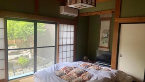 A traditional Japanese-style room with tatami flooring, a bed with floral bedding, a television, and a sliding door leading to an outdoor area with a view of greenery.