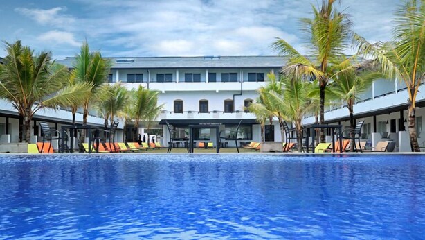 A swimming pool with lounge chairs and palm trees, surrounded by a building with multiple windows and balconies.