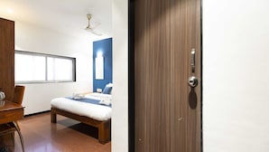 A hotel room with a bed, a desk, a chair, a window, and a door. - Hotel Q Deck Rooms (Mumbai)