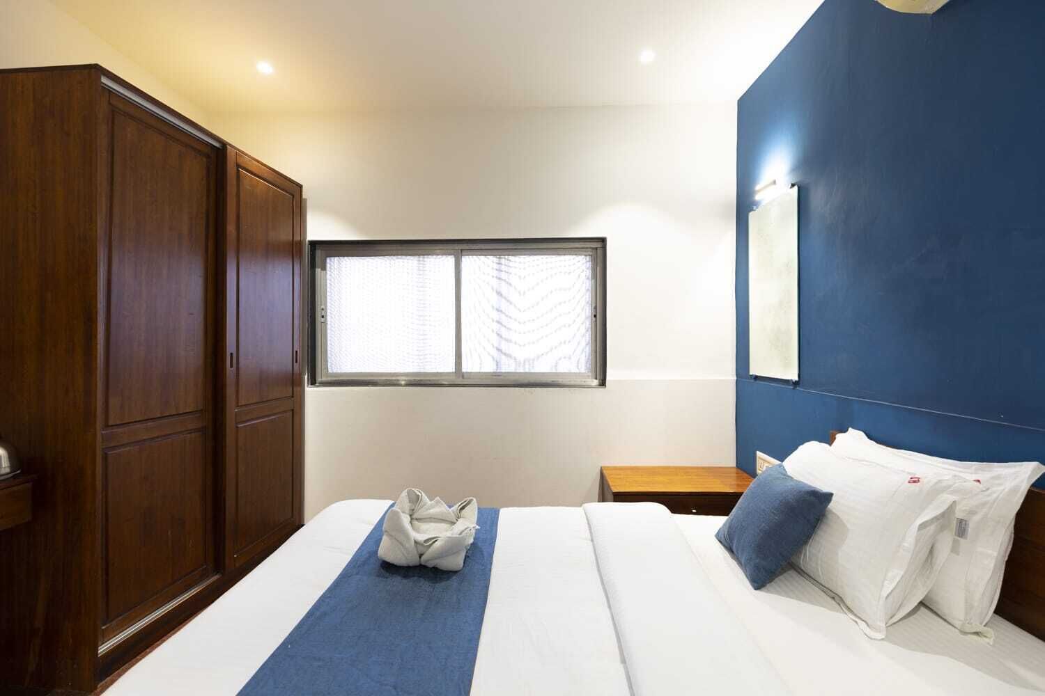 A hotel room with a large wardrobe, a bed with a blue bedspread, white pillows, and a window with blinds.