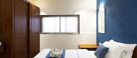 A hotel room with a large wardrobe, a bed with a blue bedspread, white pillows, and a window with blinds.