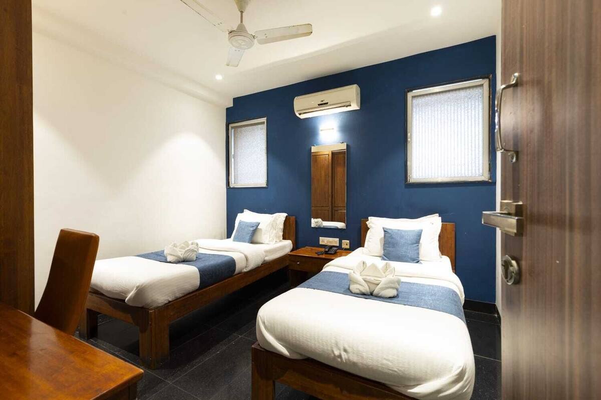 A hotel room with two single beds, a wooden desk, a chair, a ceiling fan, and an air conditioning unit.