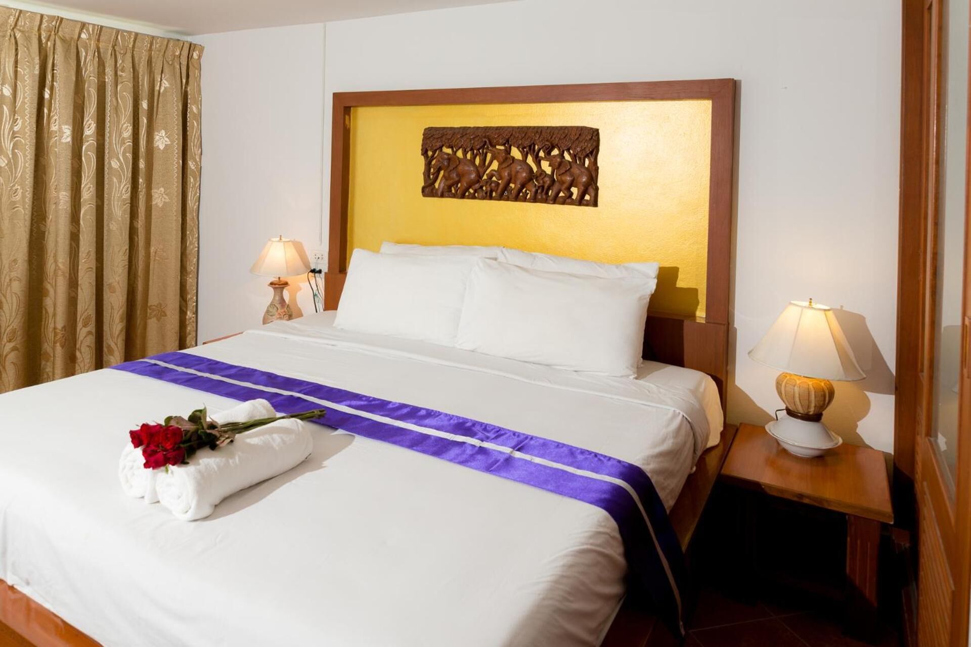 A hotel room with a large bed, a headboard with a decorative panel, two bedside lamps, and a window with curtains.