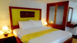 Een net opgemaakt bed met witte lakens en een gele plaids, een houten hoofdbord, een nachtkastje met een lamp en een spiegel aan de muur.