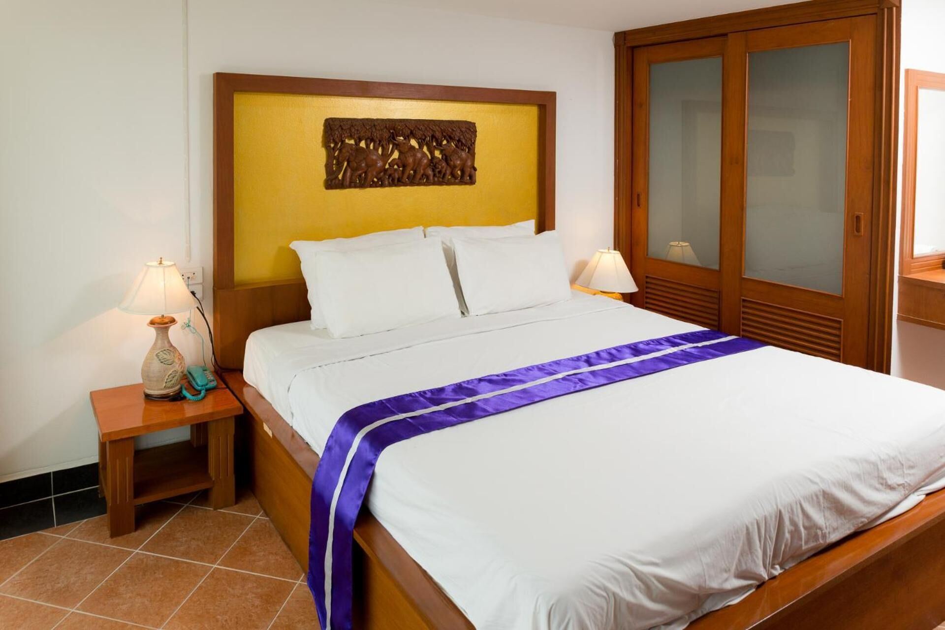 A hotel room with a wooden headboard, a bed with white linens, a purple bedspread, two bedside lamps, and a nightstand with a lamp and a telephone.