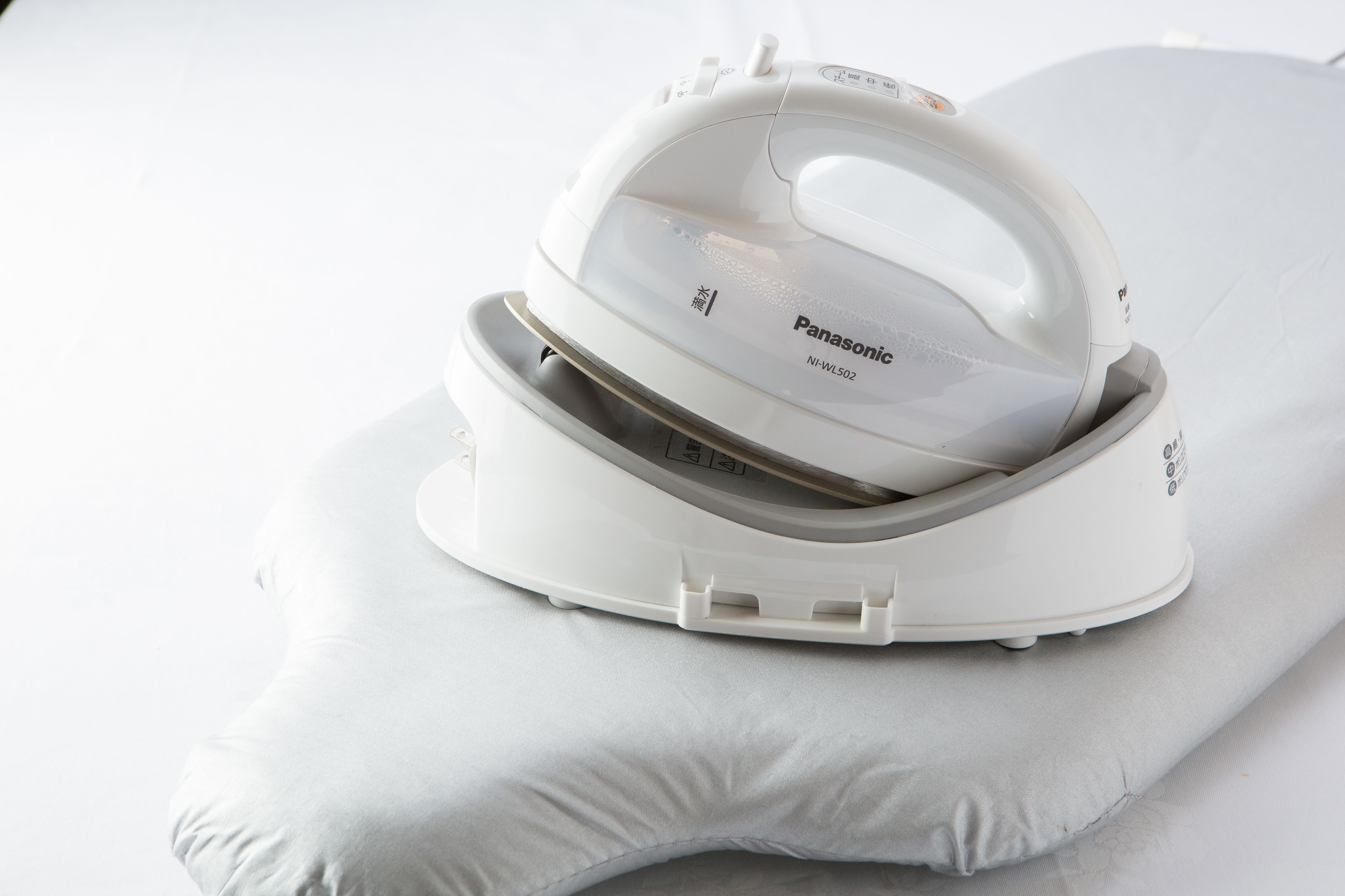 A white Panasonic iron with a transparent lid, placed on a white pillow.