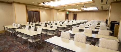 A conference room with multiple tables and chairs, arranged in rows, with a carpeted floor and a high ceiling with skylights.