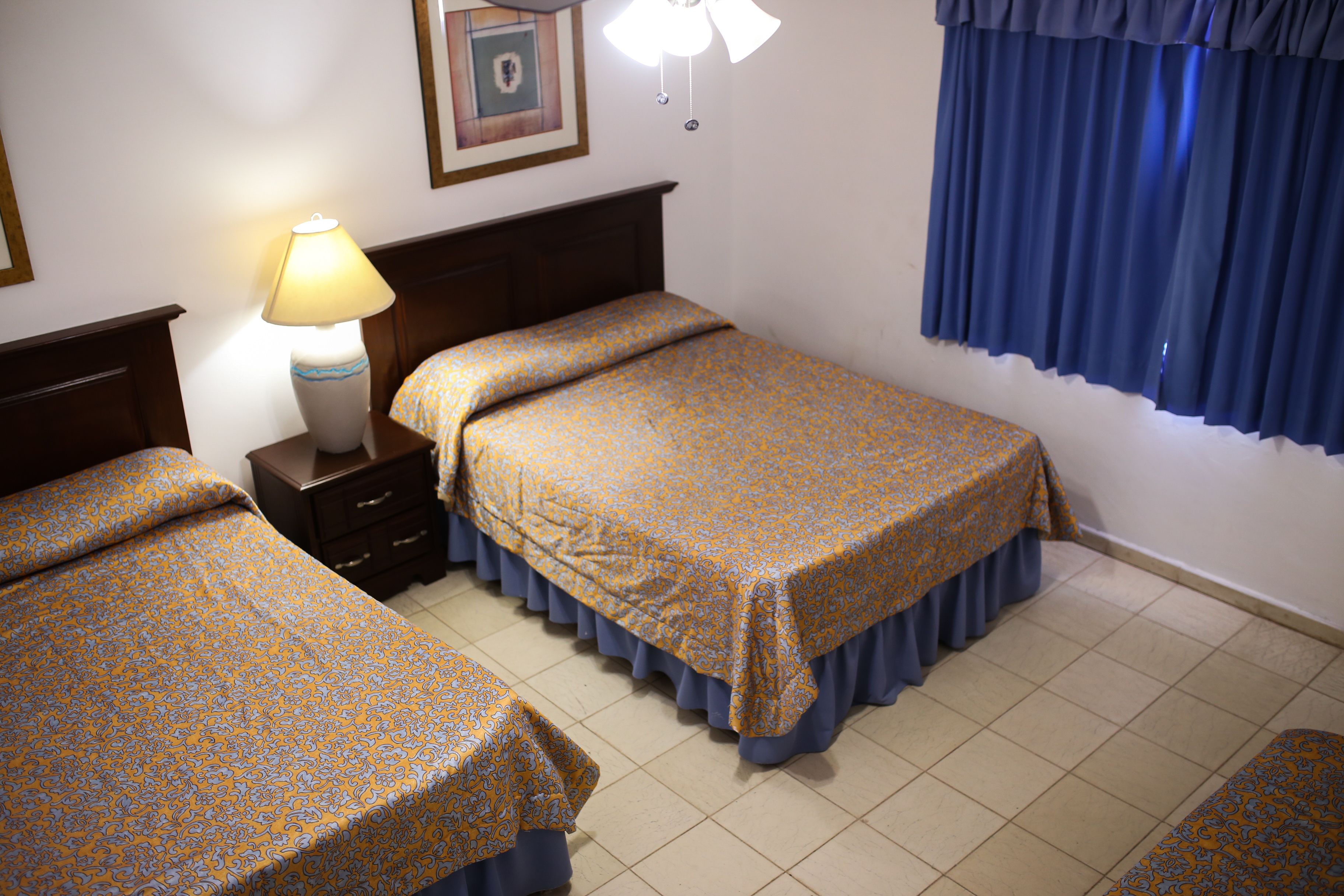 Une chambre d’hôtel avec deux lits, une table de chevet, une lampe, un tableau encadré au mur et une fenêtre avec des rideaux bleus.