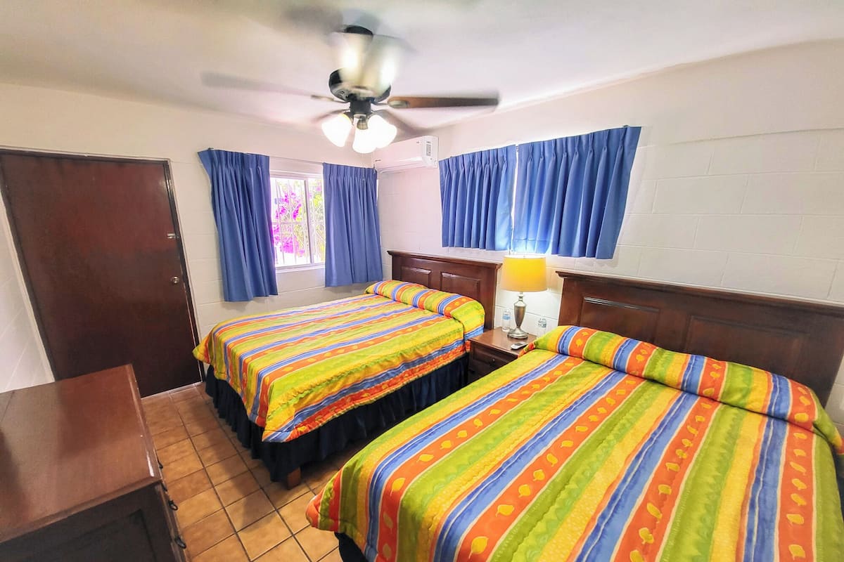 Habitación de hotel con dos camas, un ventilador de techo, una mesita de noche con lámpara y una ventana con cortinas.