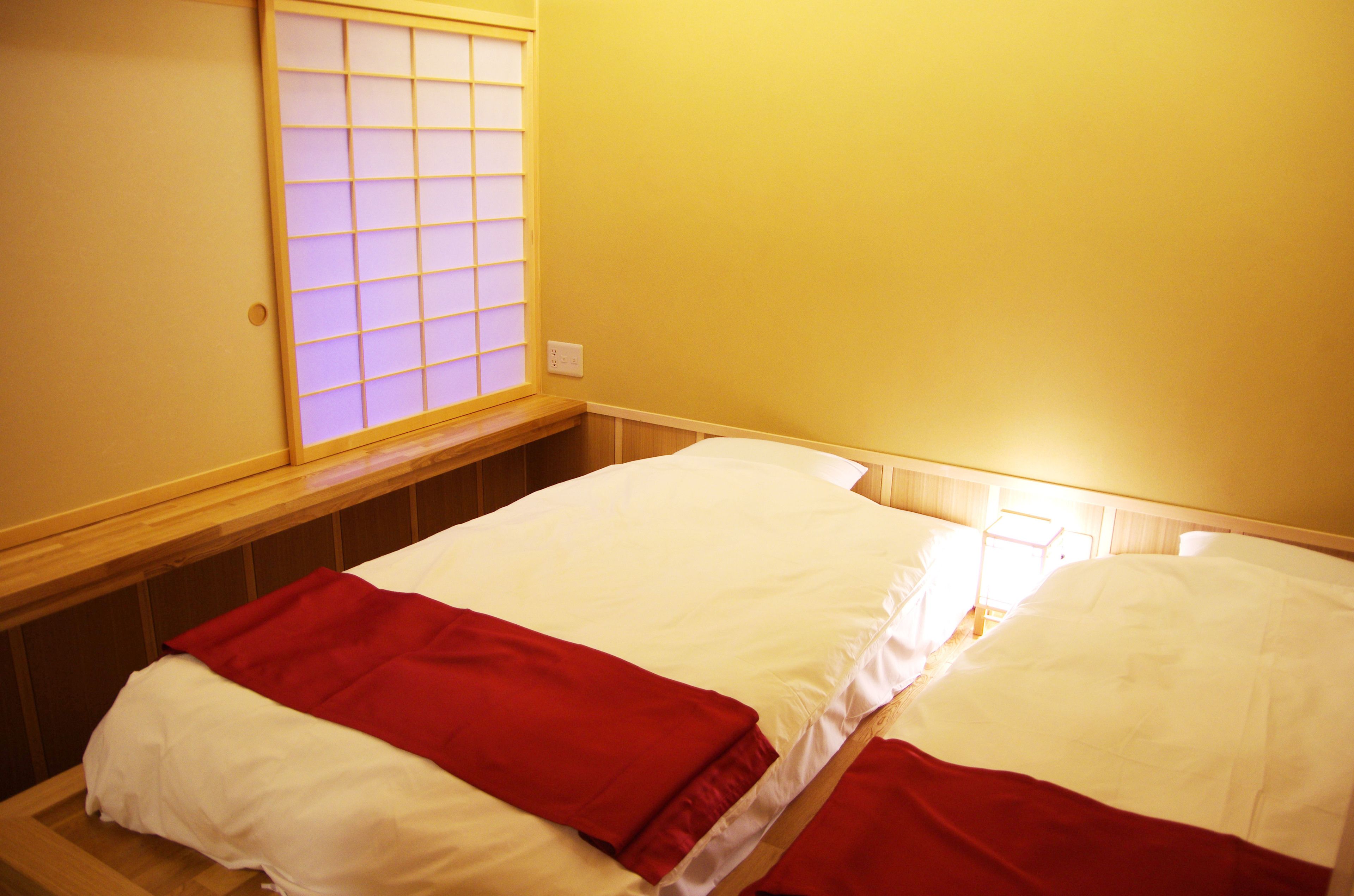 Une chambre avec deux lits, chacun doté de draps blancs et rouges, d’une tête de lit en bois, d’une fenêtre à carreaux et d’une prise de courant au mur.
