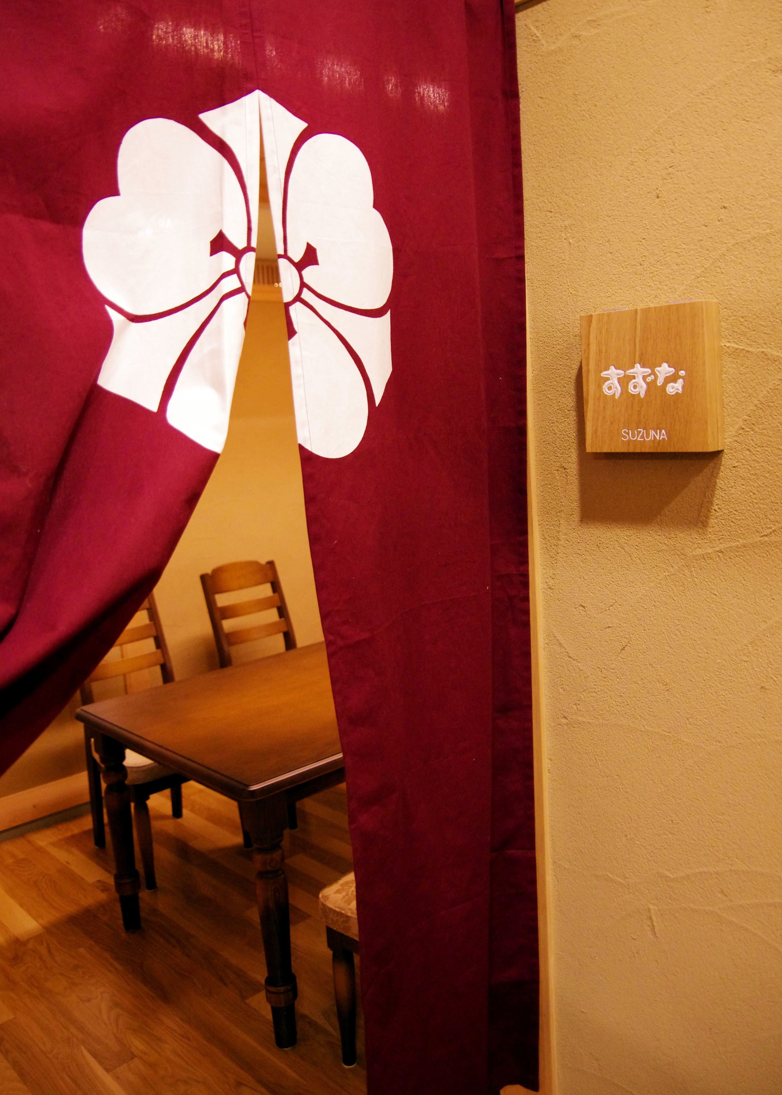 Una habitación con una mesa y sillas de madera, una cortina roja con un diseño blanco y un letrero de madera con caracteres japoneses.