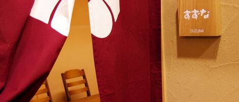 Una habitación con una mesa y sillas de madera, una cortina roja con un diseño blanco y un letrero de madera con caracteres japoneses.