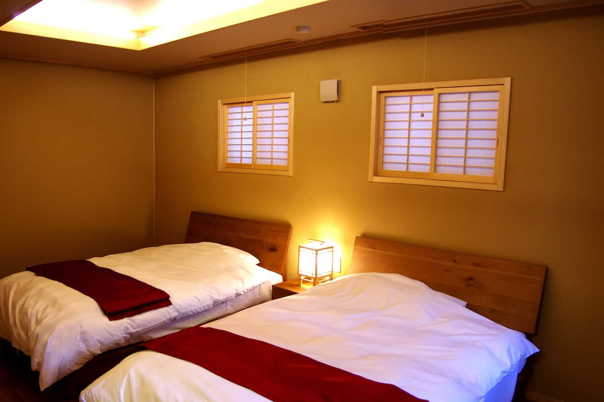 Une chambre avec deux lits, des têtes de lit en bois, une petite table avec une lampe et deux fenêtres équipées de stores.