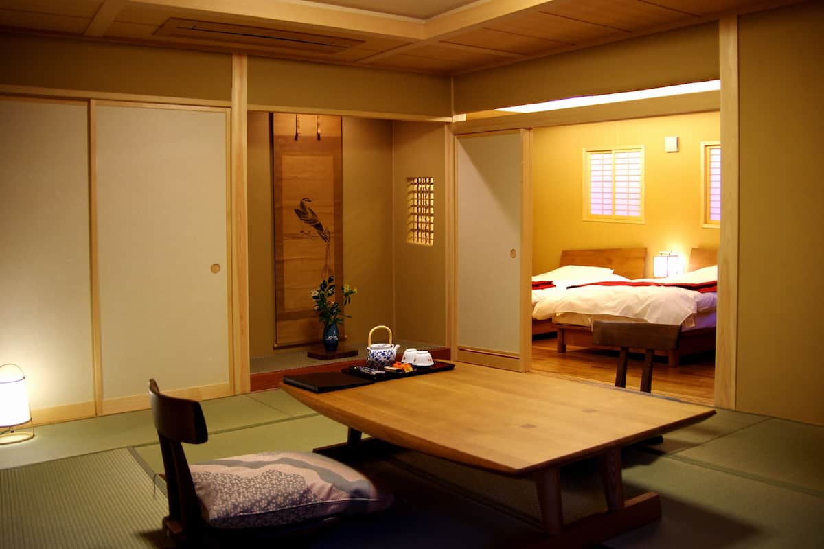 Une pièce de style japonais traditionnel, avec un sol recouvert de tatamis, une petite table en bois, une chaise et un lit doté d’une tête de lit en bois.