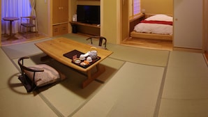 Une pièce de style japonais traditionnel, avec un sol recouvert de tatamis, une petite table en bois sur laquelle est disposé un service à thé, un lit recouvert d’une couverture rouge et un téléviseur posé sur une étagère basse.