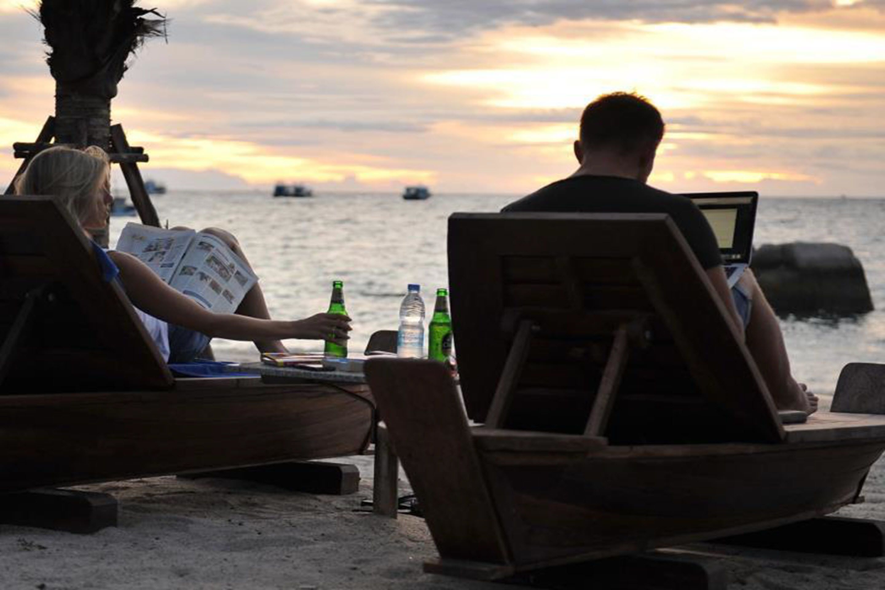 Two people relaxing on wooden lounge chairs by the beach with a laptop and drinks, overlooking the ocean.