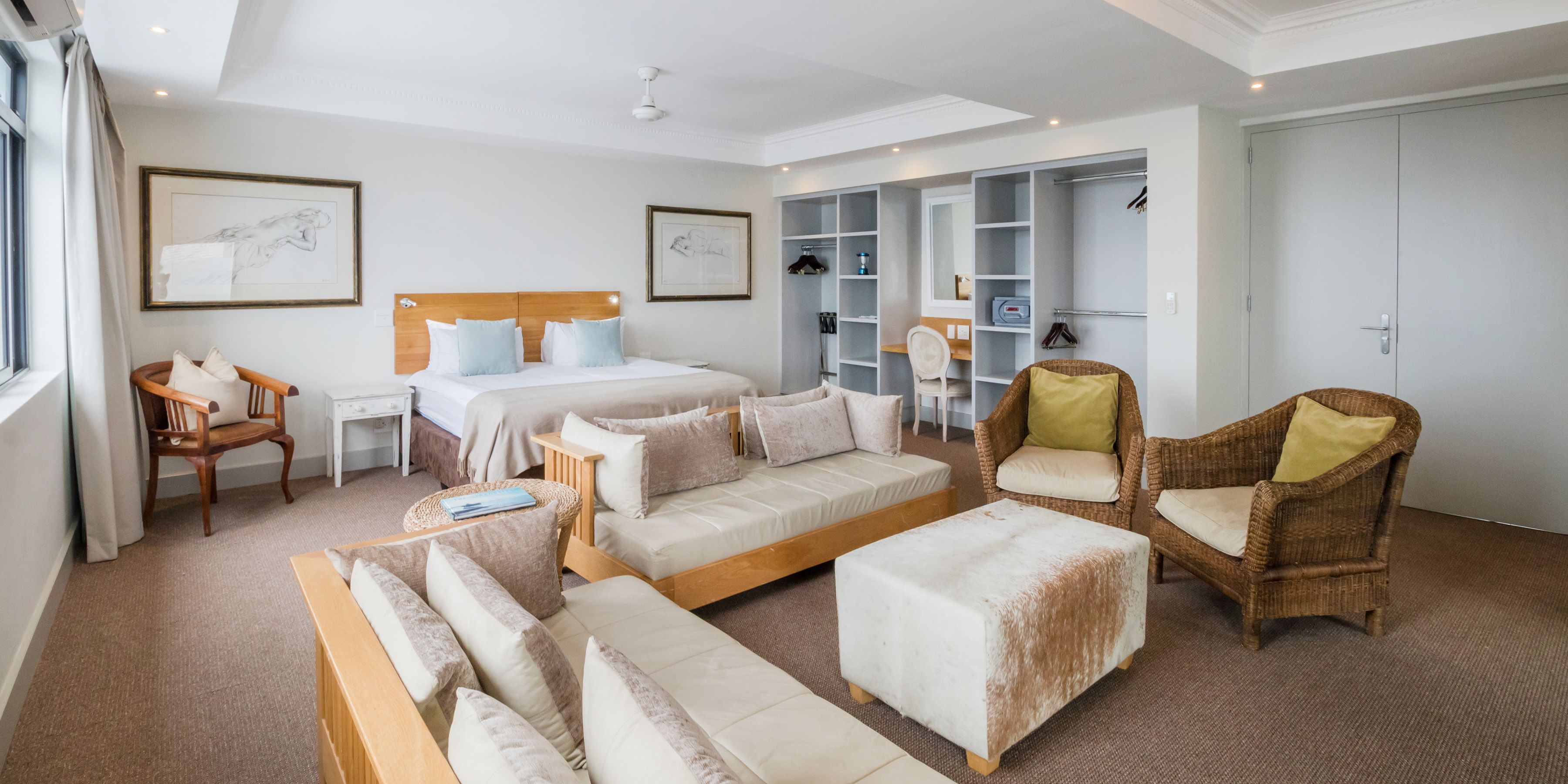 A modern hotel room with a large bed, two sofas, a small table, wicker chairs, and built-in wardrobes.