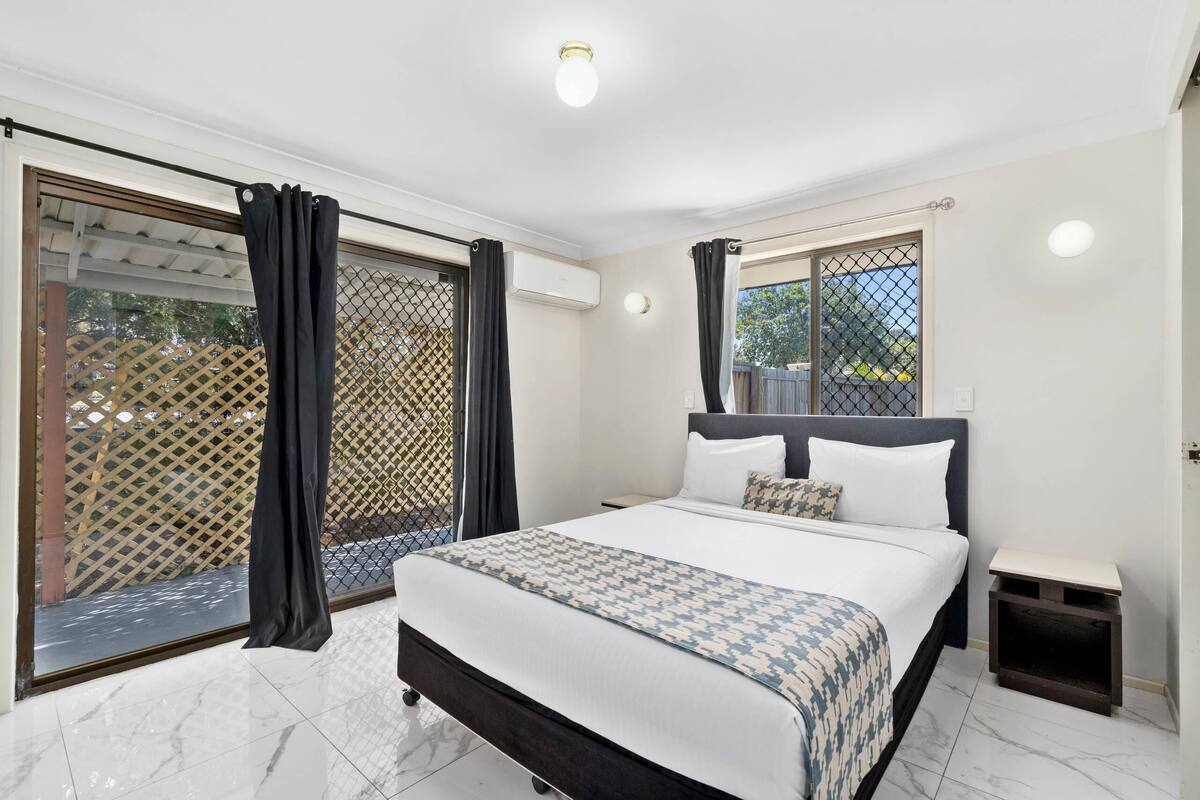 A bedroom with a bed, a nightstand, a window with curtains, and a view of the outside.