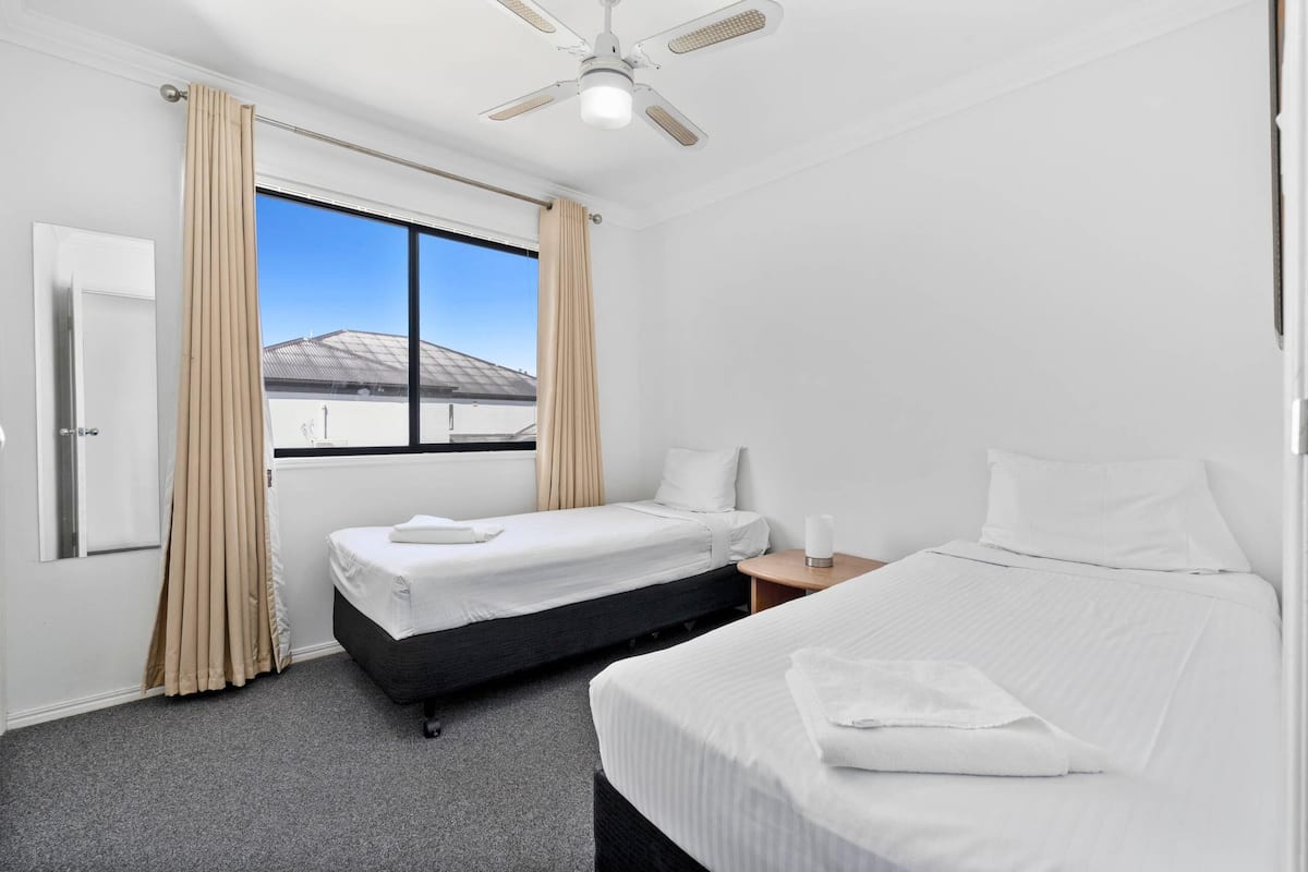 A hotel room with two beds, a window with curtains, a ceiling fan, and a view of a building outside.