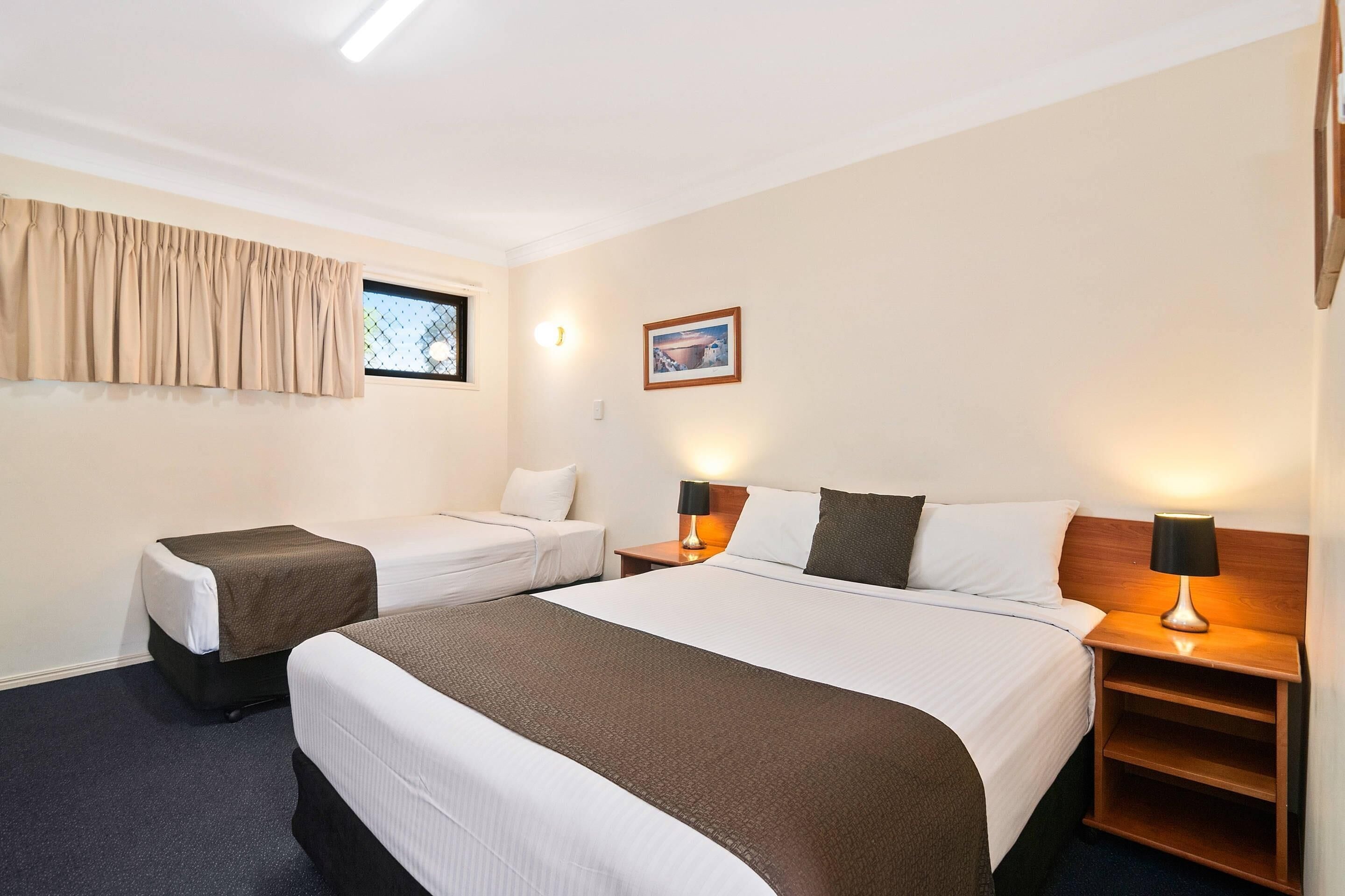 A hotel room with two beds, a window with curtains, a nightstand with a lamp, and a framed picture on the wall.