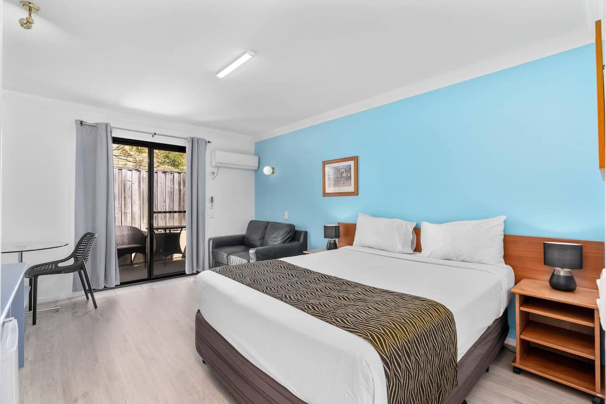 A hotel room with a bed, a desk, a chair, a nightstand, and a view of the outside.