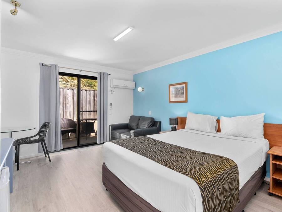 A hotel room with a bed, a desk, a chair, a nightstand, and a view of the outside.