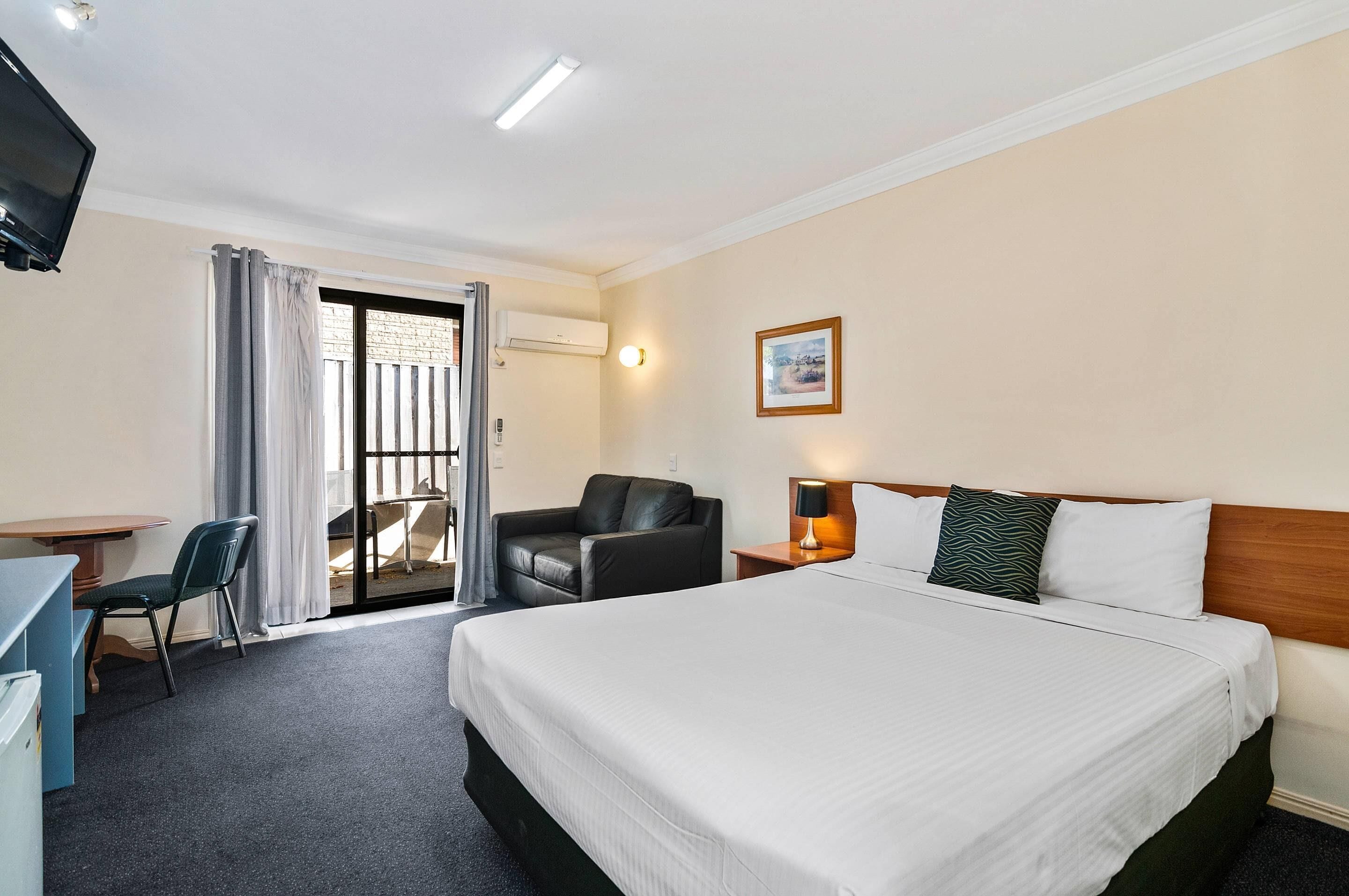 A hotel room with a bed, a desk, a chair, a TV, a window with curtains, and a wall-mounted lamp.