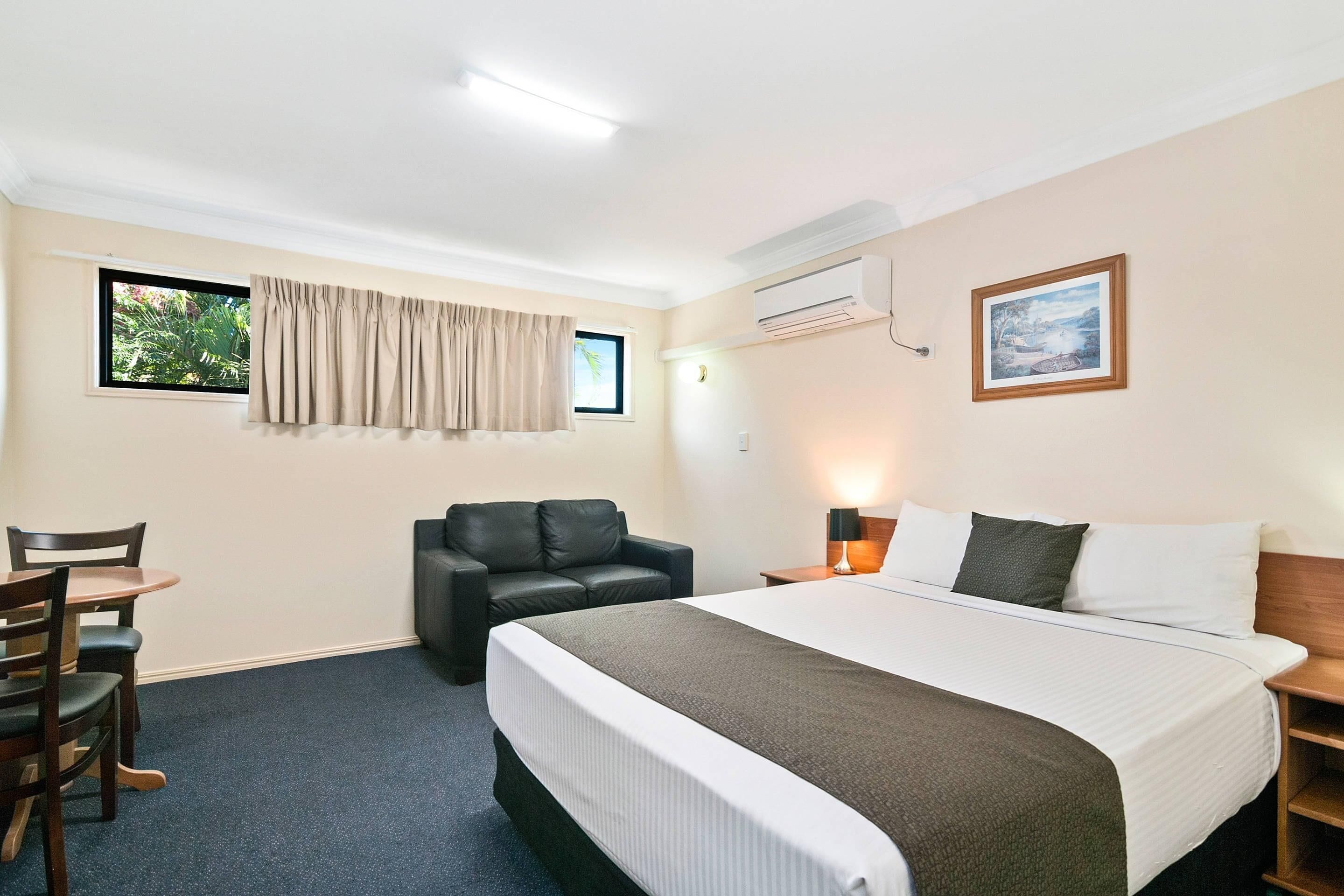 Habitación de hotel con cama, escritorio con sillas, sofá de cuero, ventana con cortinas, equipo de aire acondicionado y un cuadro en la pared.
