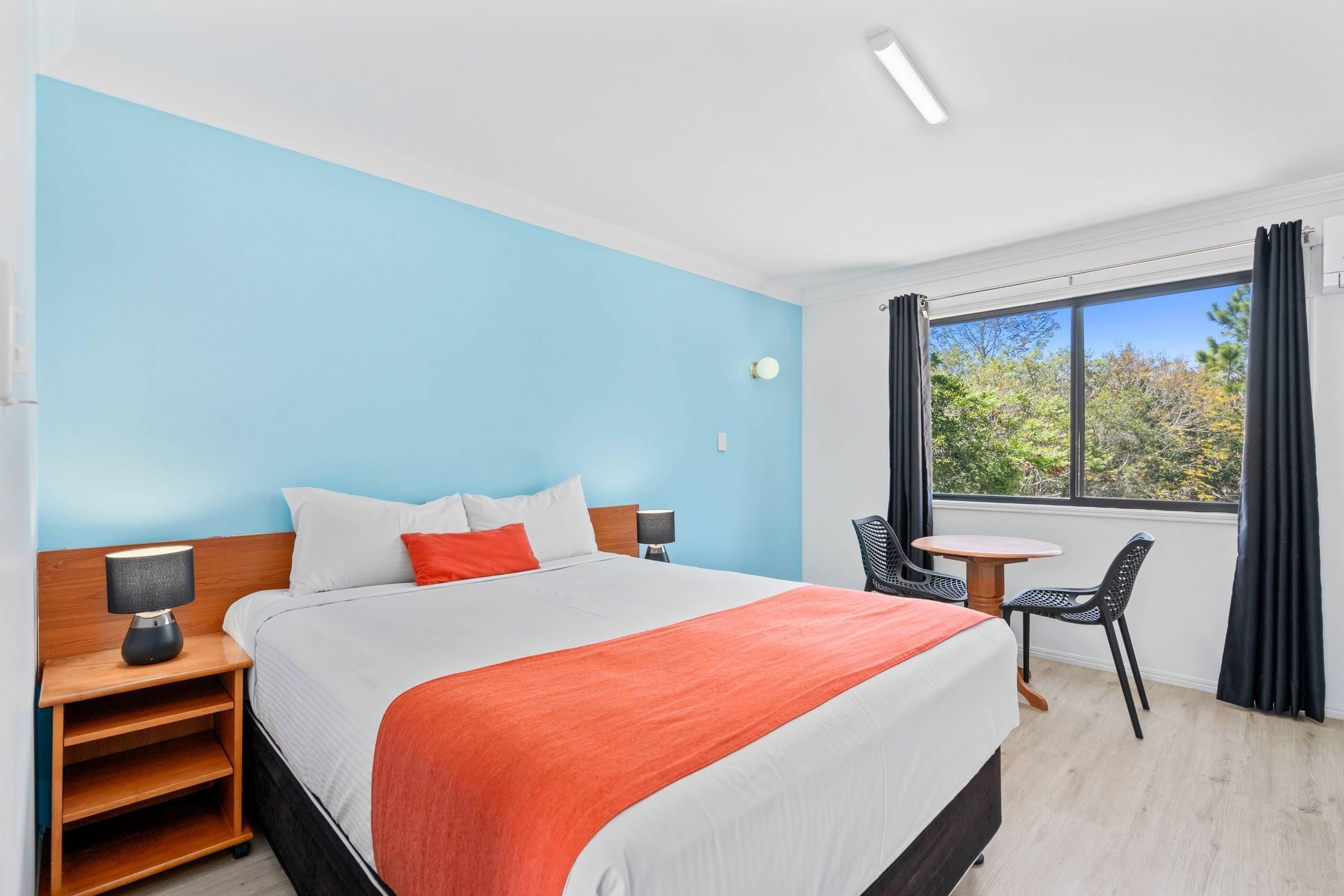 A hotel room with a bed, bedside tables, a chair, a small table, and a window with curtains.
