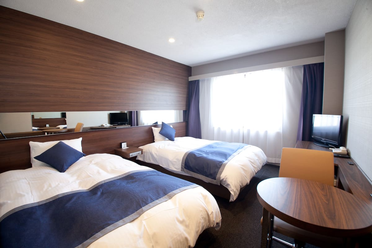 Une chambre d’hôtel avec deux lits, un bureau, une chaise, une télévision et une fenêtre avec des rideaux.