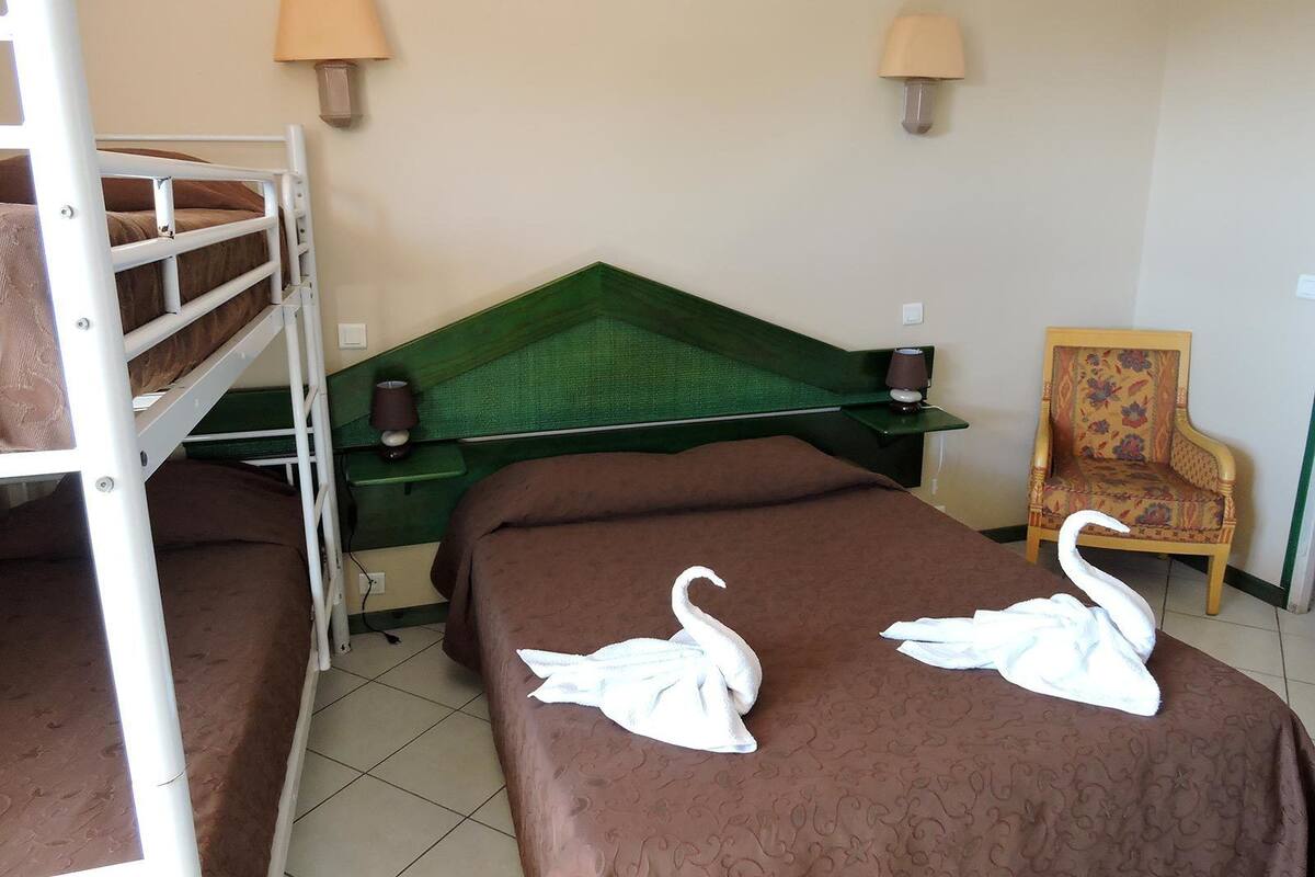Une chambre d’hôtel avec deux lits simples, une tête de lit verte, deux lampes murales, un fauteuil à motifs floraux et deux serviettes en forme de cygne posées sur les lits.