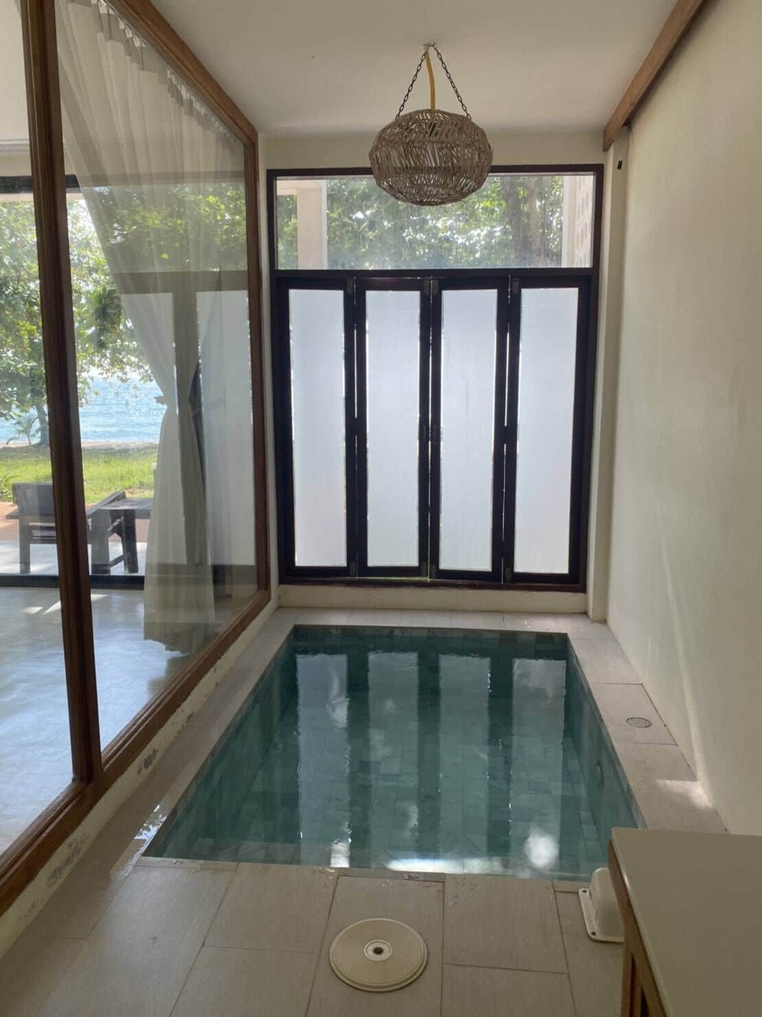 Una piscina interior pequeña con paredes de vidrio transparente, una lámpara en el techo y vistas al exterior.