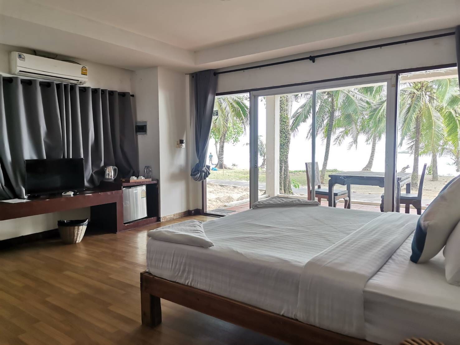 A bedroom with a bed, a TV, a desk, and a large window with a view of palm trees and beach.
