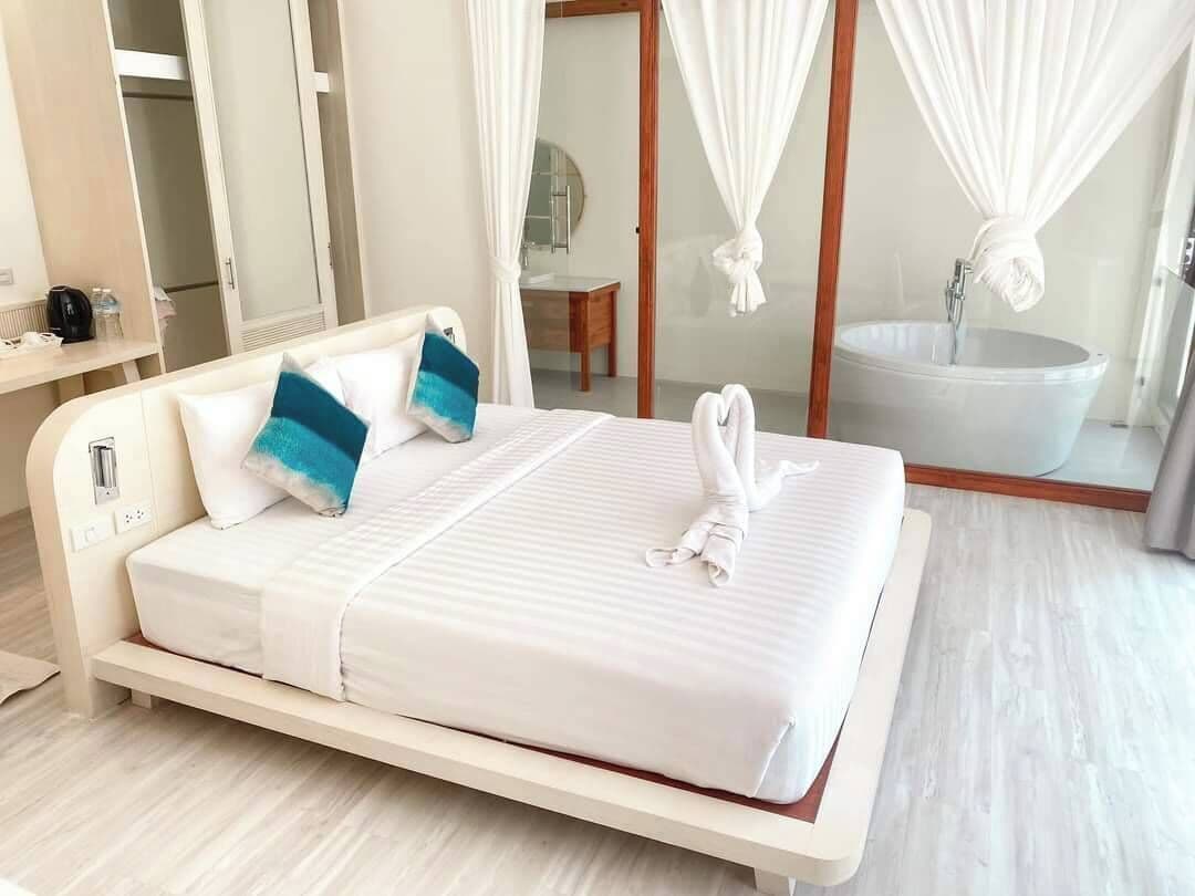 Een net opgemaakt bed met wit beddengoed en blauwe kussens, een nachtkastje met een lamp, en een badkamer met een bad en een handdoek aan de douchestang.