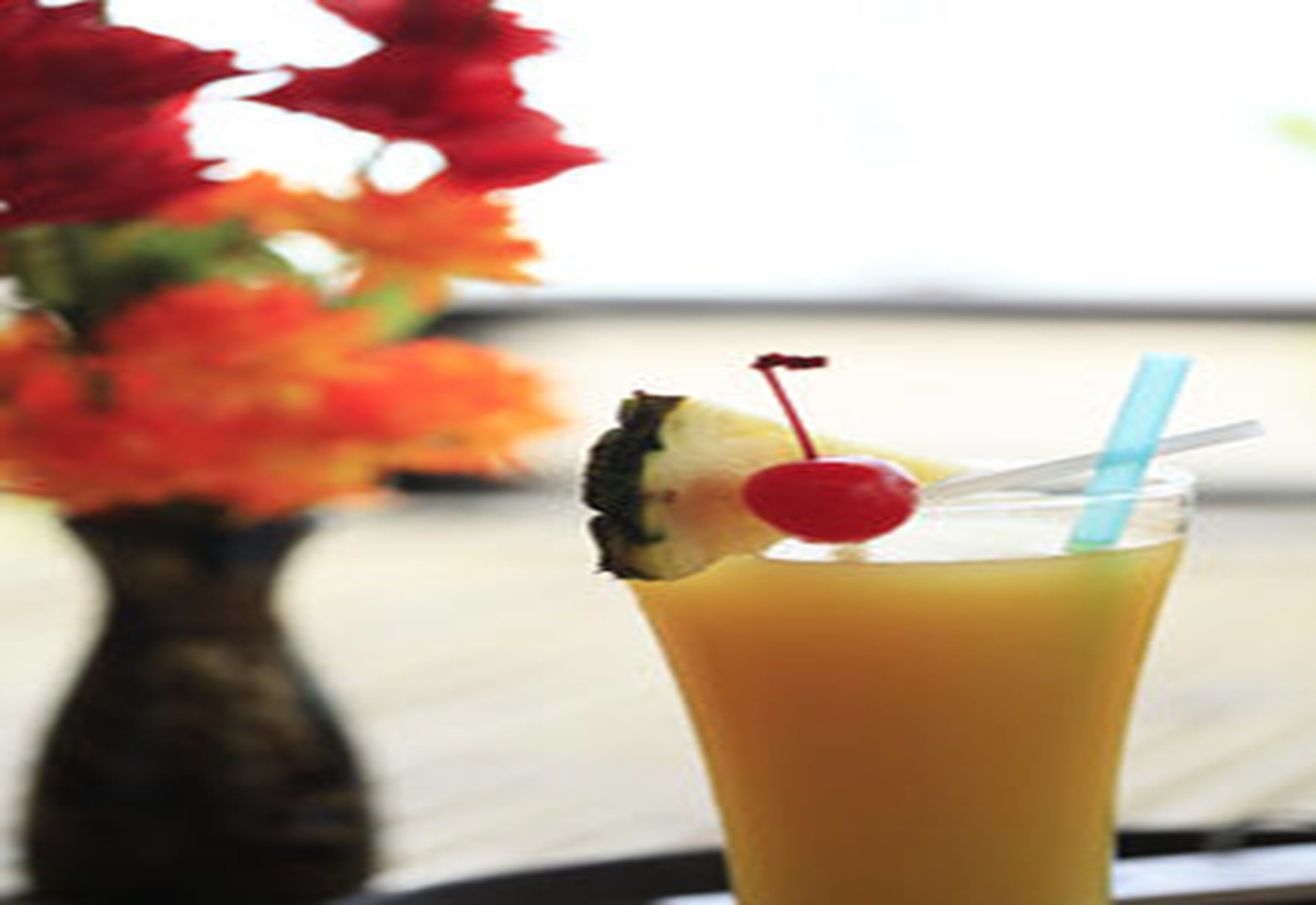 A tall glass with a yellow beverage, a cherry, and a straw.