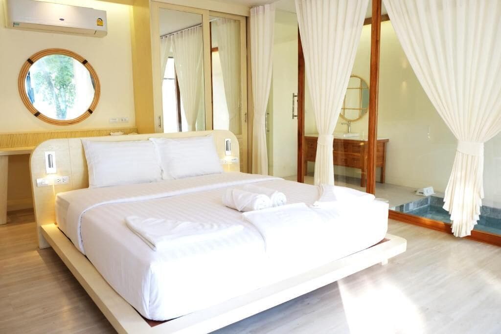 A large bed with white linens, a bedside table with a lamp, a round mirror, and a view of a pool through a glass door.