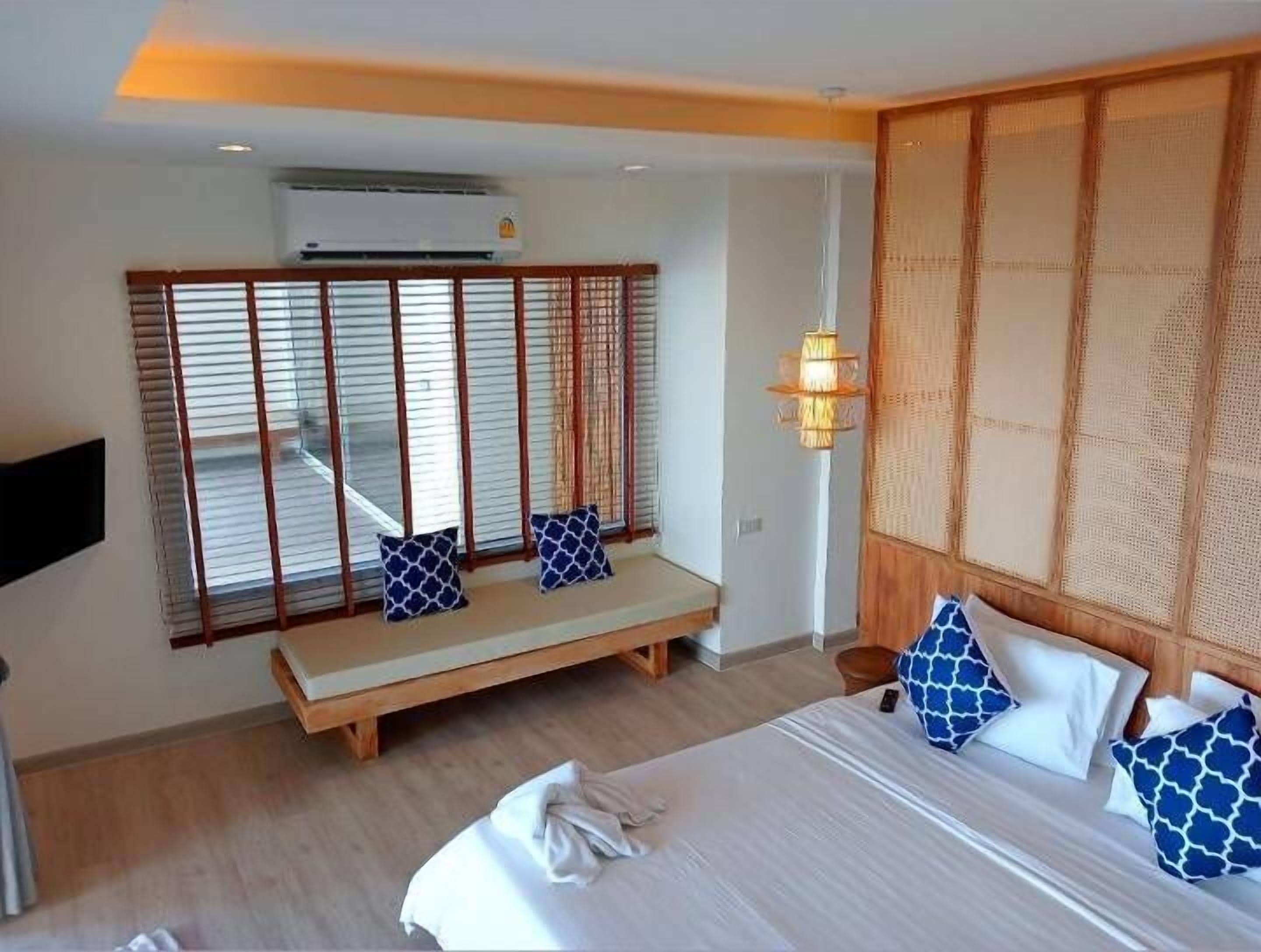 A hotel room with a bed, a bench with pillows, a wall-mounted TV, and a large window with blinds.